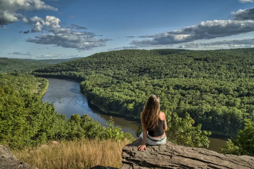 Girl sitting on a mountain ledge in the Catskills, enjoying the breathtaking views of the river and surrounding landscape in spring.