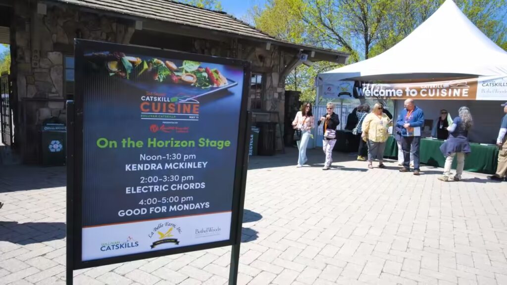 People enjoying the Catskills Cuisine event with a vendor tent and sign board in the background, showcasing local food and vendors at the event.